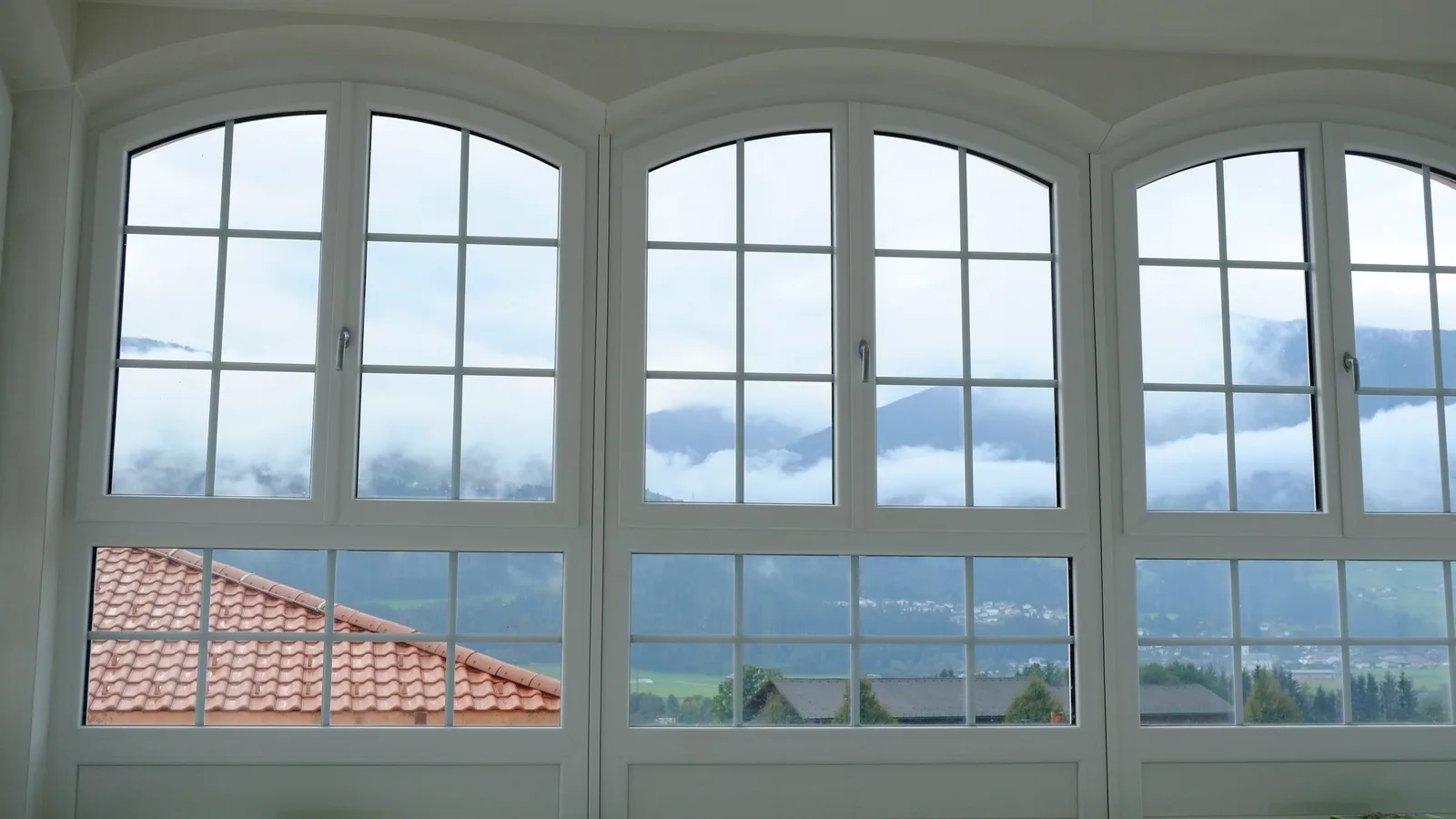 Eine Fensterfront aus weißen Rundbogenfenstern in einem Rohbau, dahinter Nebel und Dächer.