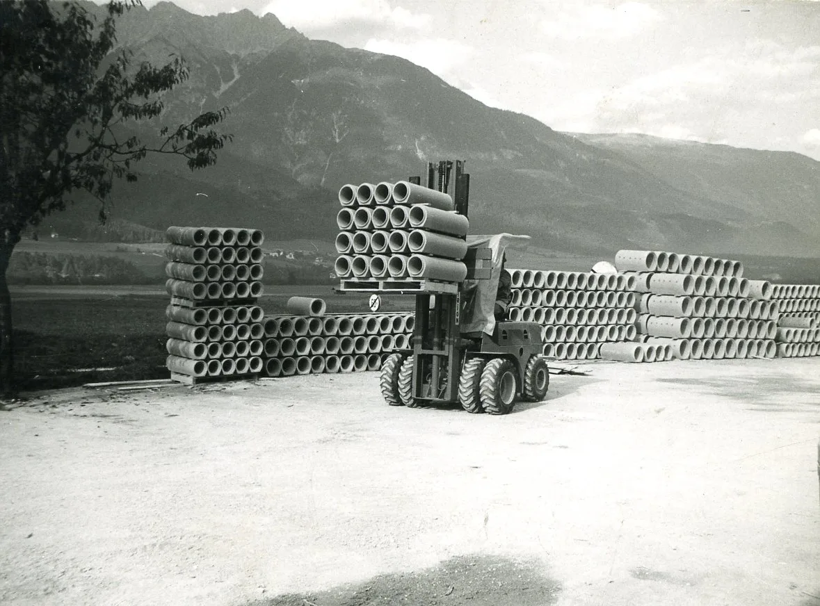 Schwarzweißaufnahme von 1966: ein Stapler zwischen hohen Stapeln aus Betonrohren, dahinter die Berge.