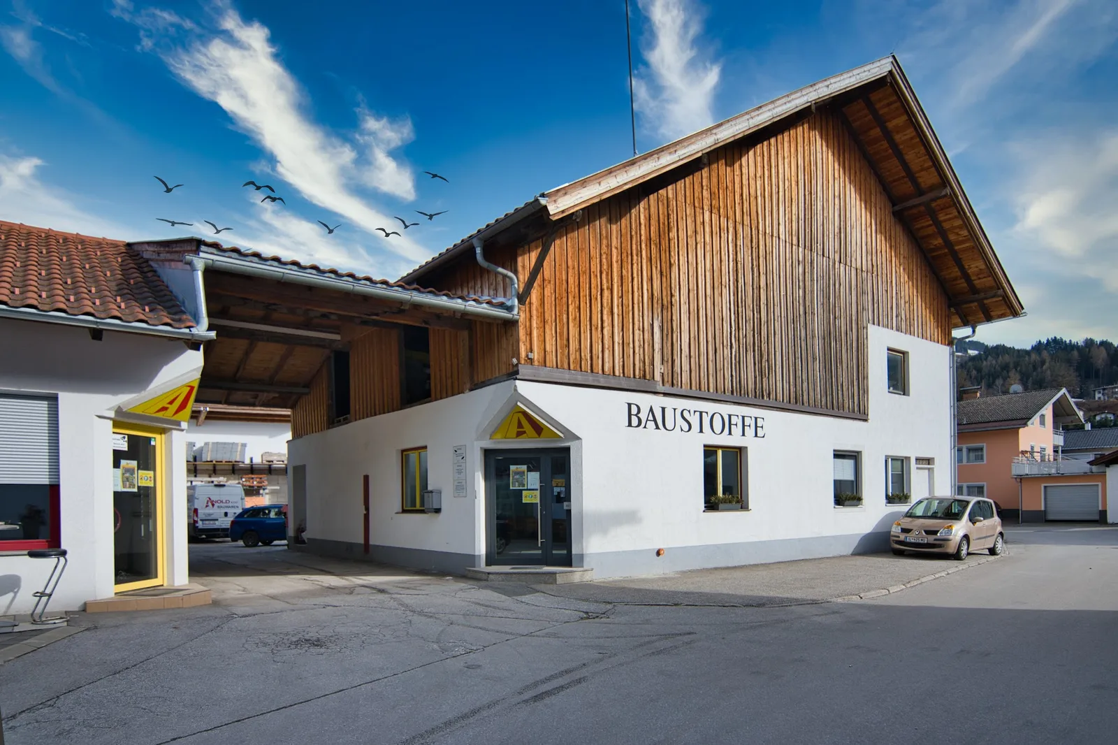 Das Firmengebäude in Weer: weiße Wand mit dem Schriftzug Baustoffe, darüber eine Holzfassade, über der Eingangstür das gelbe A-Zeichen.