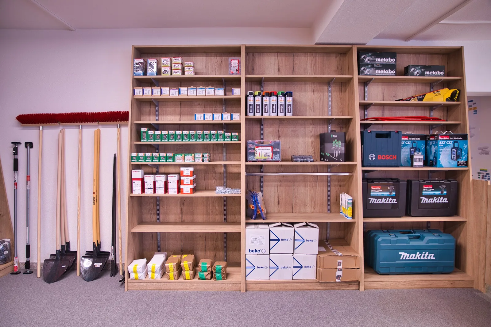 Regalwand im Verkaufsraum mit Schraubenschachteln, Dübeln, Sprühdosen, Elektrowerkzeug in Koffern sowie Schaufeln und Besen an der Wand.