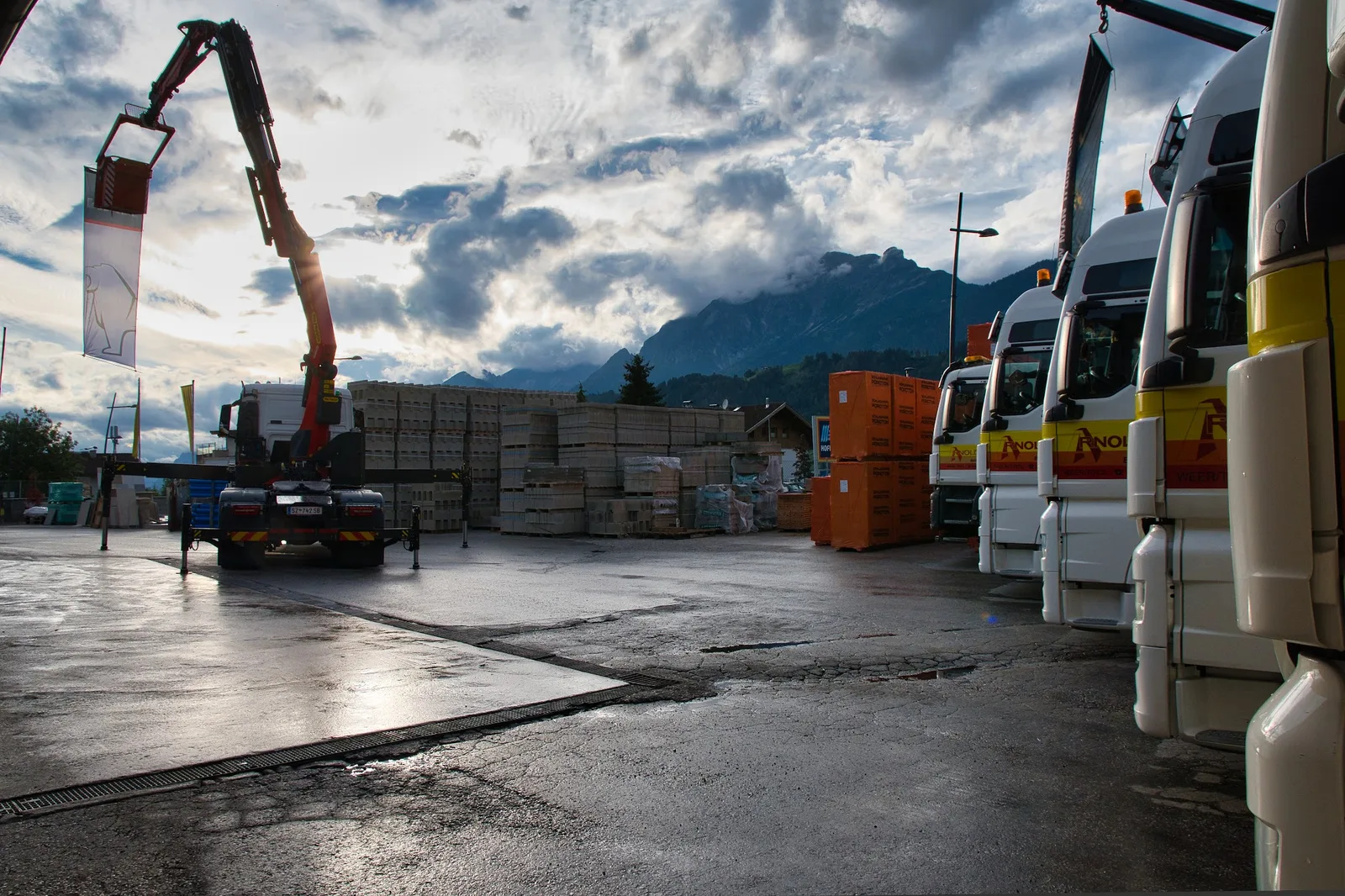 Der Lagerplatz in der Dämmerung nach Regen, eine Reihe beschrifteter Lastwagen, dahinter Betonwaren und die Berge.