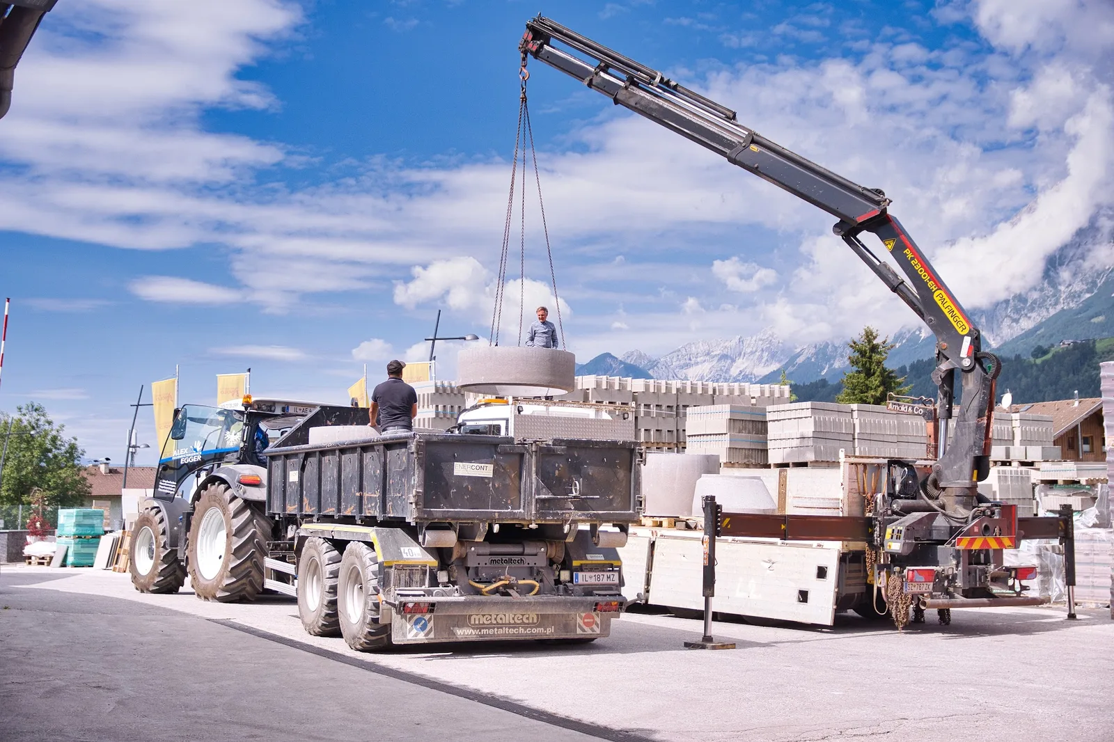 Ein Ladekran hebt einen Betonring auf einen Anhänger, daneben stehen Betonwaren und Randsteine, im Hintergrund die Berge.